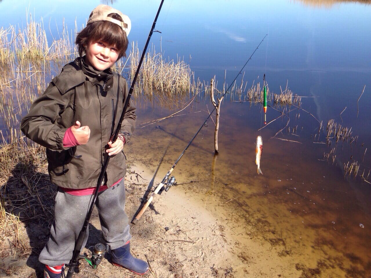 Ein Junge mit dunkler Jacke, Kappe und Gummistiefeln steht am Ufer und hält eine Angelrute mit kleinem Fisch, Schilf und Wasser im Hintergrund.
