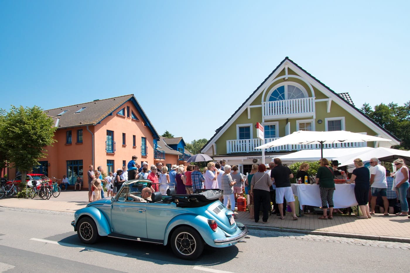 Ein hellblaues Cabriolet fährt an einer Menschenmenge vor dem Café Pieni in Ahrenshoop vorbei.