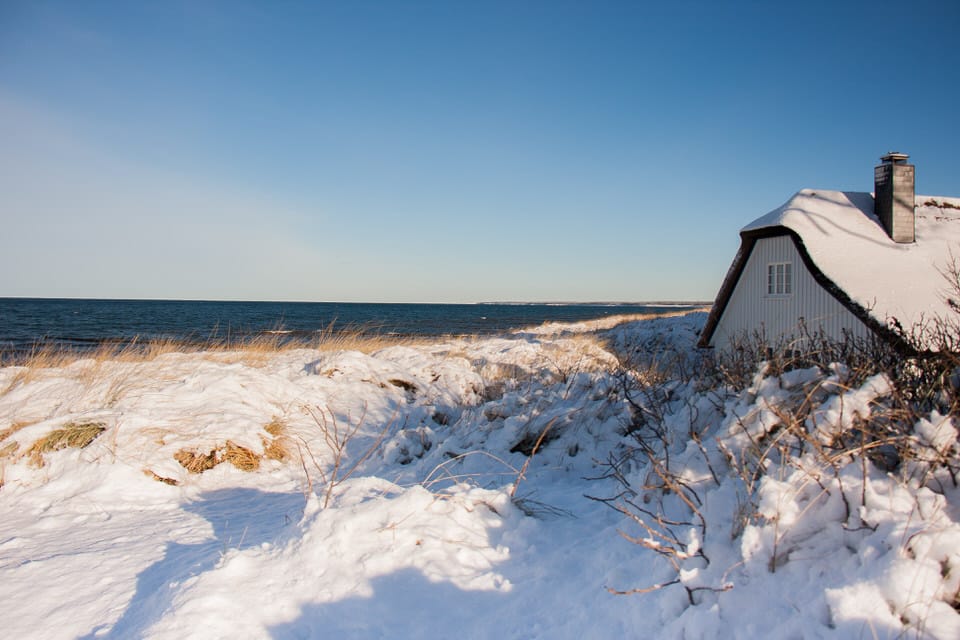 Ein Haus steht am Rand der verschneiten Dünen. Im Hintergrund das Meer und ein klarer Himmel.