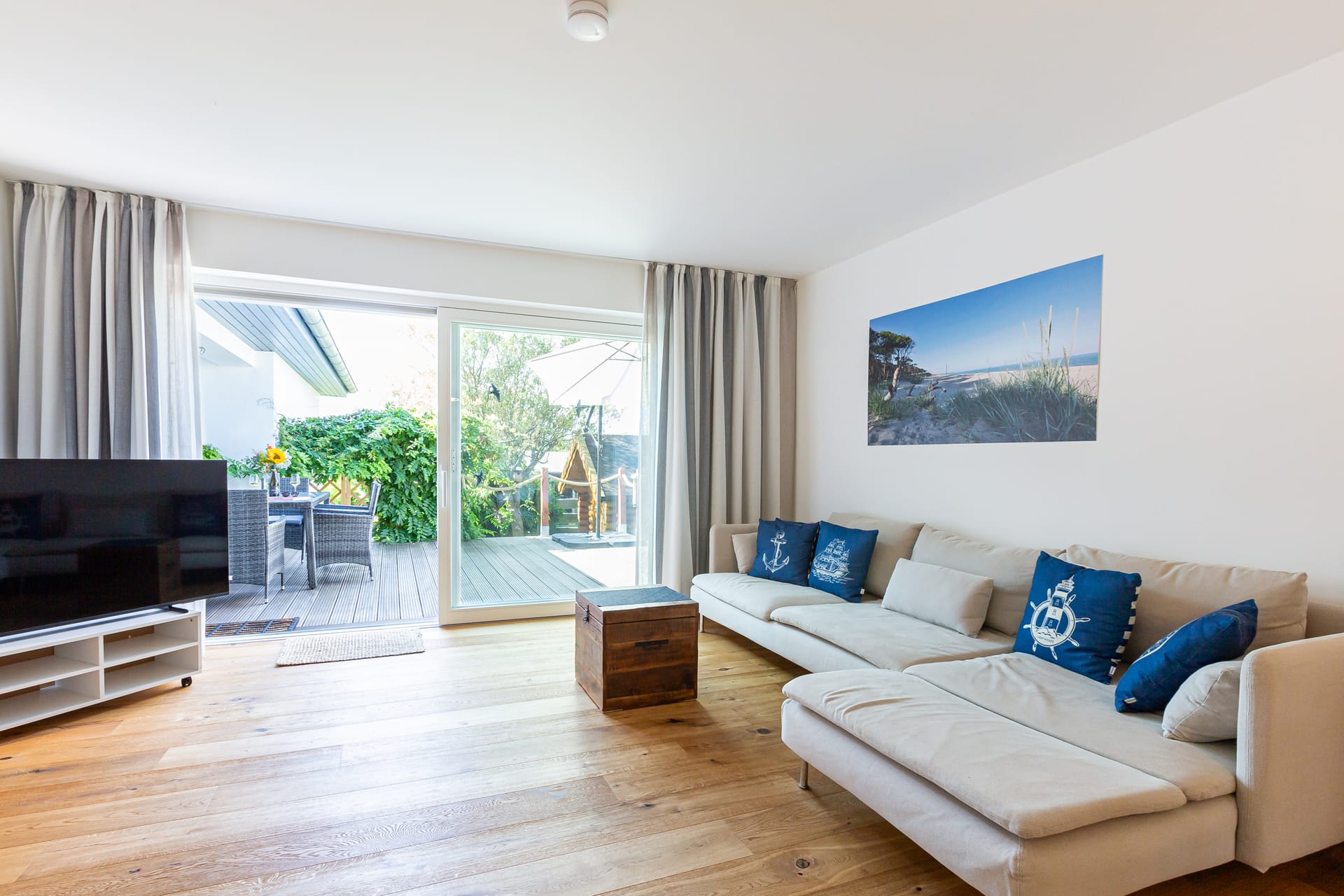 Im Wohnzimmer steht links der Fernseher, rechts an der Wand eine große, helle Couch mit blauen Kissen. Über der Couch hängt ein Bild. Weiter hinten führt eine Tür auf die Terrasse, auf der Gartenmöbel stehen, und dahinter sieht man üppiges Grün.