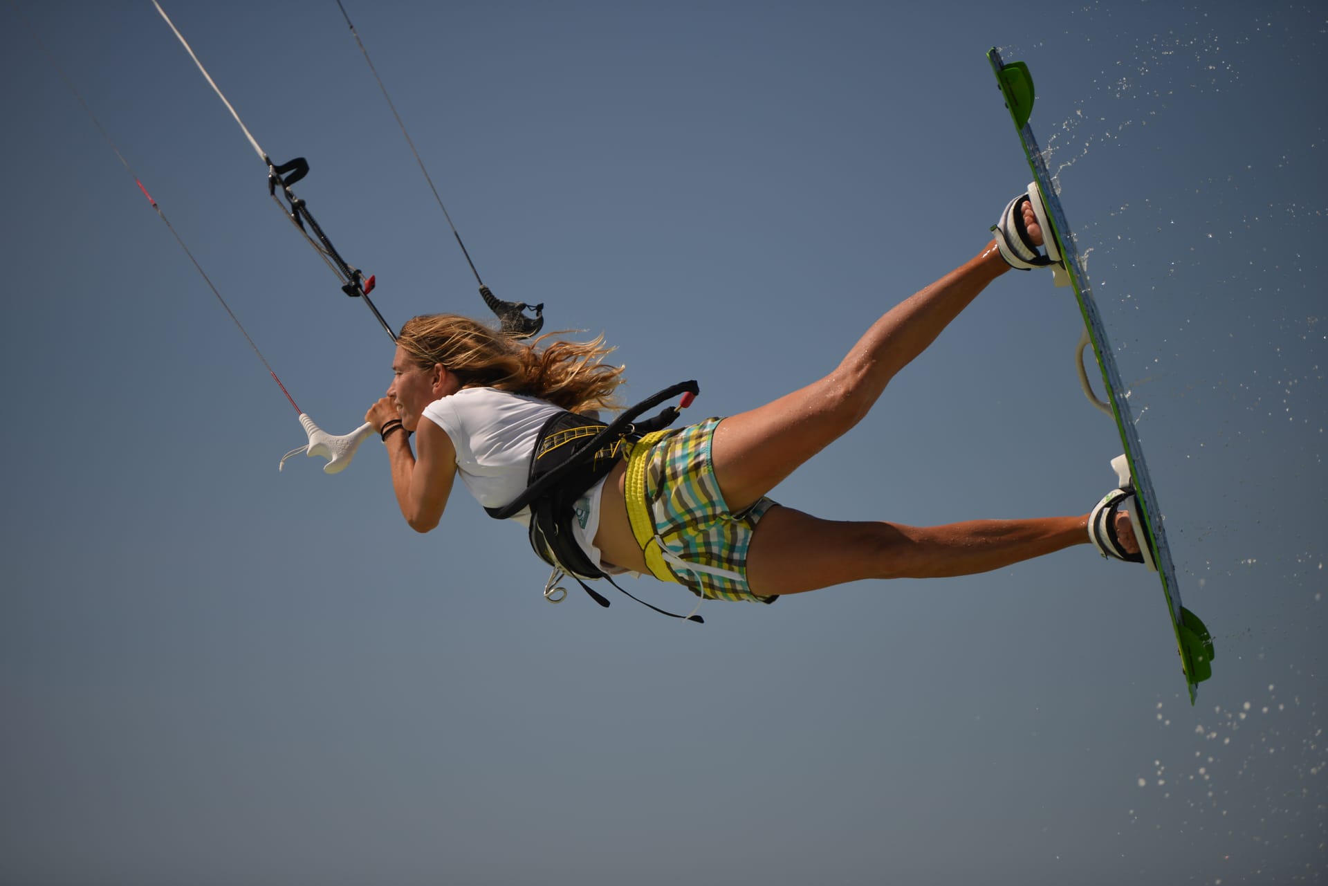 Eine Person fliegt beim Kitesurfen hoch in der Luft, an einem Kite befestigt, über dem Wasser unter blauem Himmel.
