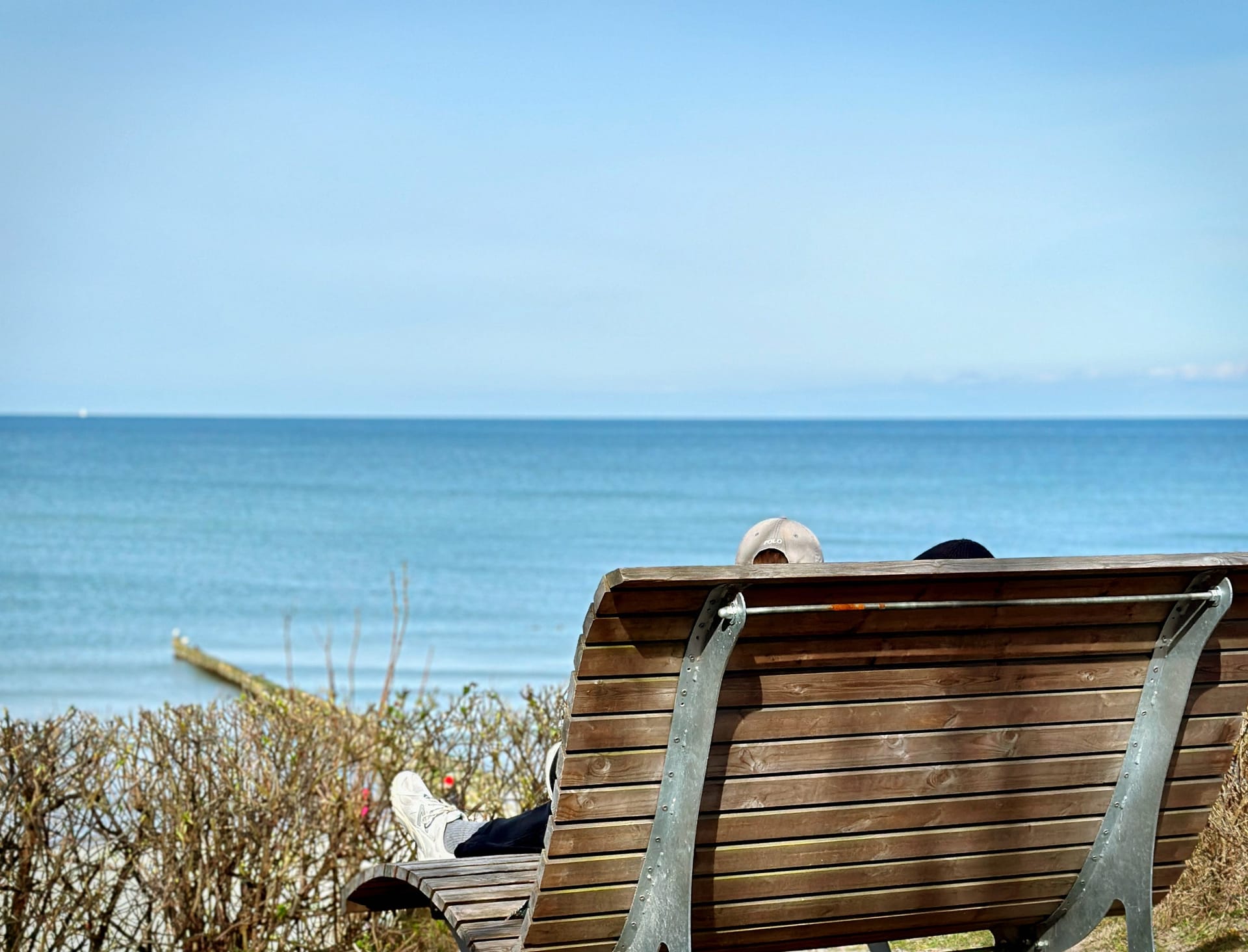 Strandkulisse - im Vordergrund ist eine Bank zu sehen, auf der Menschen sitzen. Im Hintergrund erstrecken sich das blaue Meer und der Himmel. Die Aufnahme zeigt die Strandlandschaft von oben.