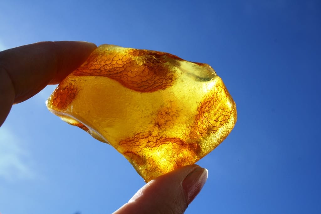 Ein großer, gelb-transparenter Bernstein mit Einschlüssen zwischen Fingern vor blauem Himmel im Sonnenlicht.