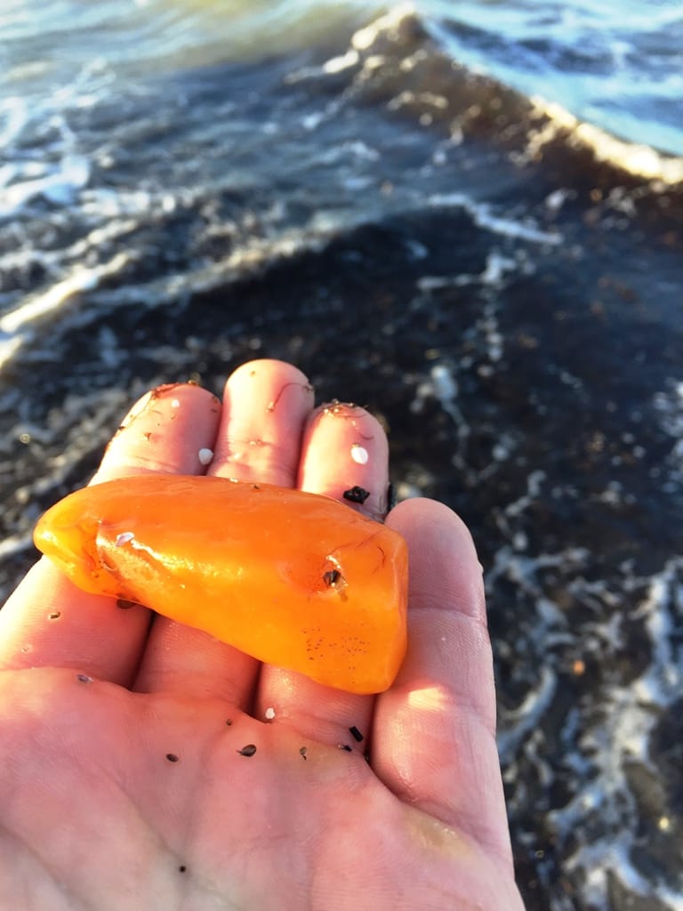 Ein großer, orangefarbener Bernstein in einer Hand am Meeresufer mit Wellen im Hintergrund.