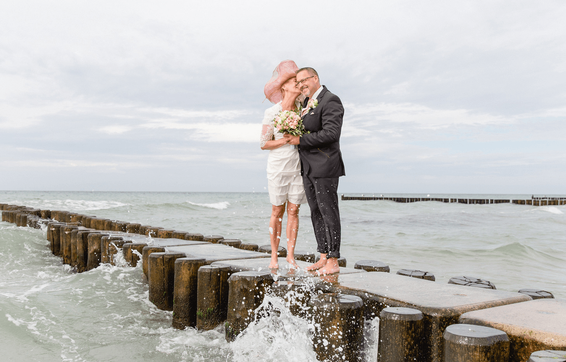 Ein Brautpaar barfuß auf einem Holzsteg im Meer, umarmt sich und hält einen Blumenstrauß, während Wellen um sie herumspritzen.