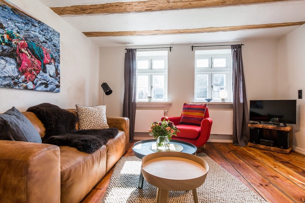 Wohnzimmer mit braunem Ledersofa, rotem Sessel und Holzbalkendecke vor zwei Fenstern.