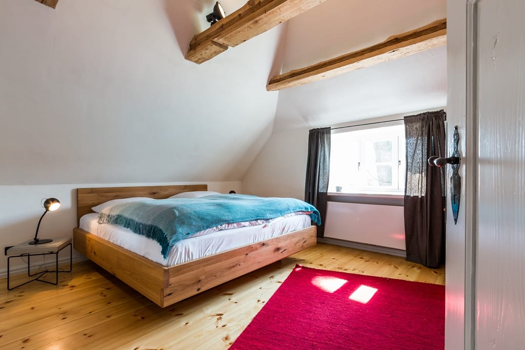 Schlafzimmer unter der Dachschräge mit Holz-Doppelbett, Balken, Dielenboden und rotem Teppich.
