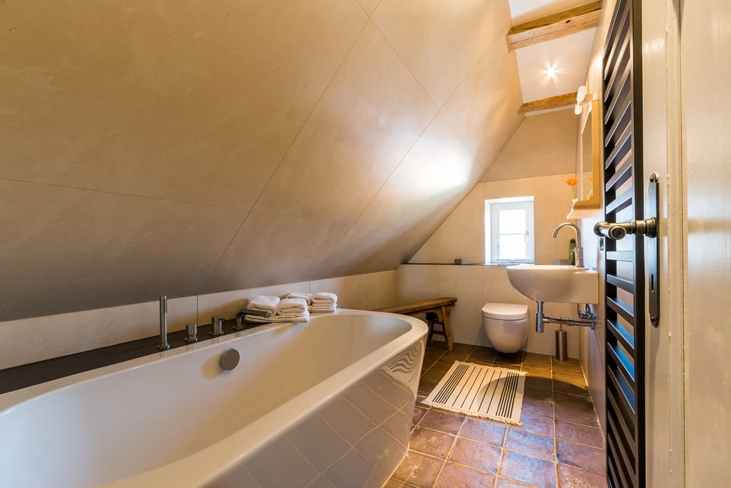 Badezimmer unter einer Dachschräge mit einer weißen Badewanne, einem Waschbecken, einem WC und braunen Bodenfliesen.