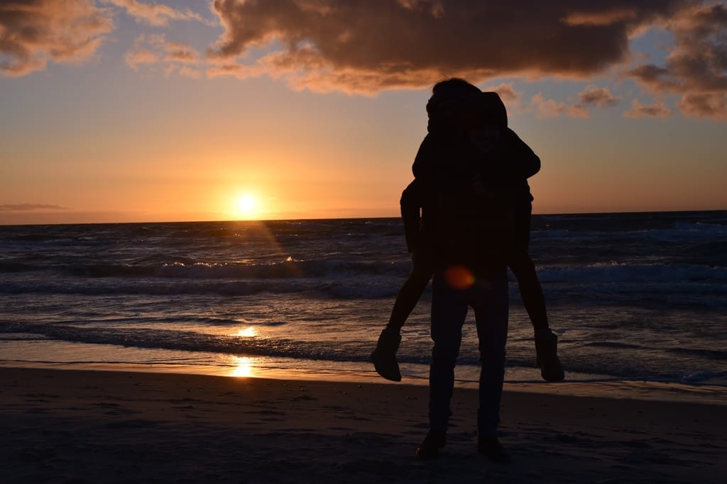 Ein Paar am Strand bei Sonnenuntergang, er trägt sie auf dem Rücken.