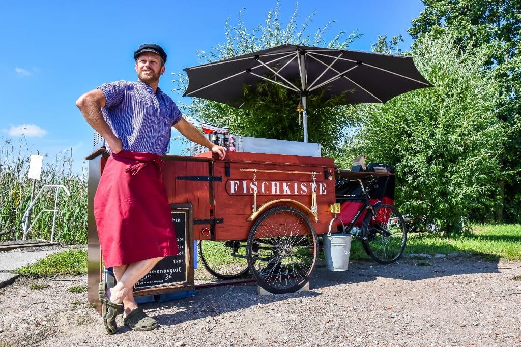 Ein Mann in Arbeitskleidung neben einem mobilen Verkaufsstand mit der Aufschrift „FISCHKISTE“ unter einem Sonnenschirm.