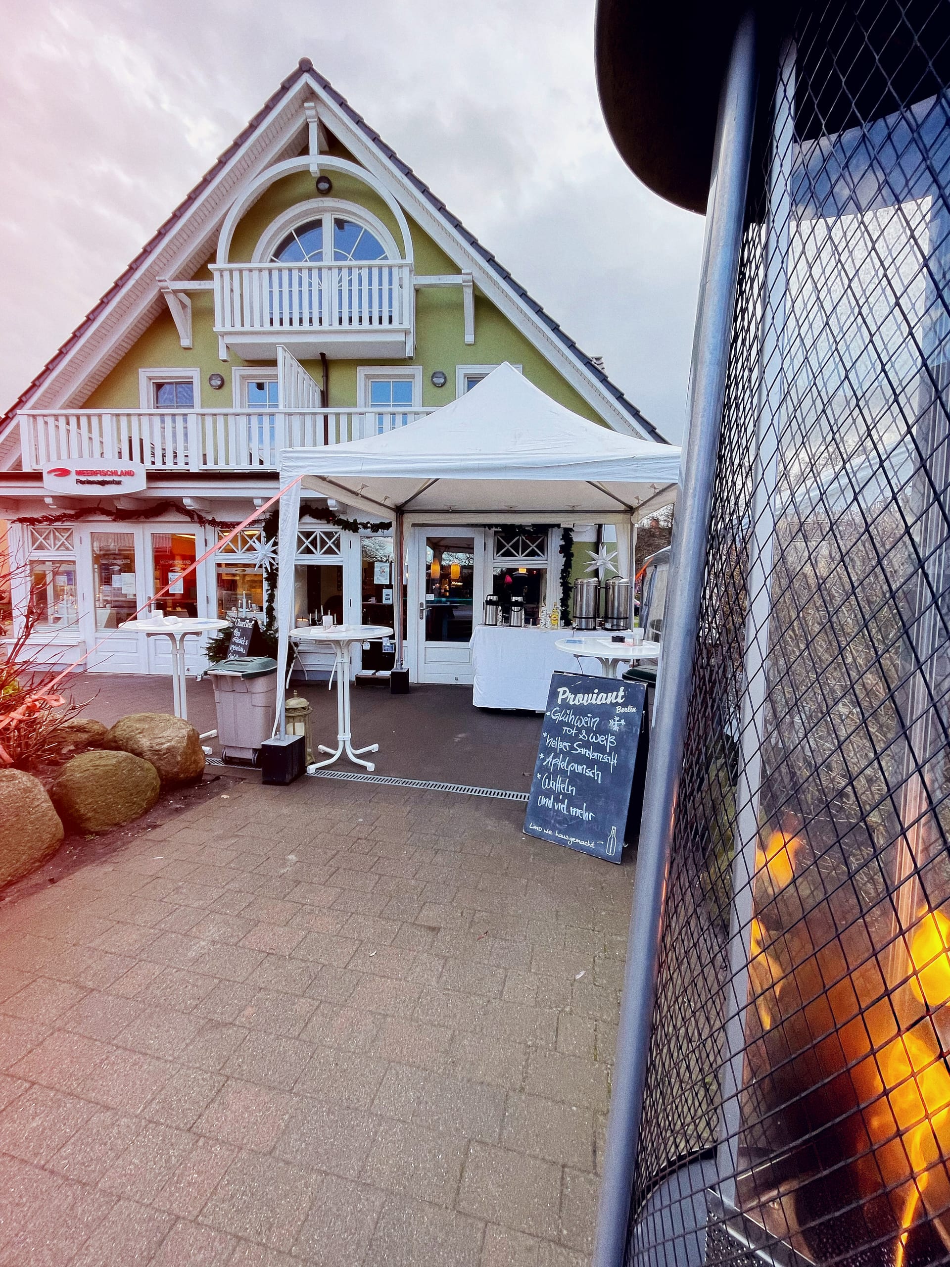 Eingang zum Café, davor ein weißes Zelt mit weißen Stehtischen. Auf den Tischen Thermoskannen mit heißen Getränken. Eingang weihnachtlich geschmückt.t. Auf dem Aufsteller werden Glühwein und heiße Getränke angeboten.