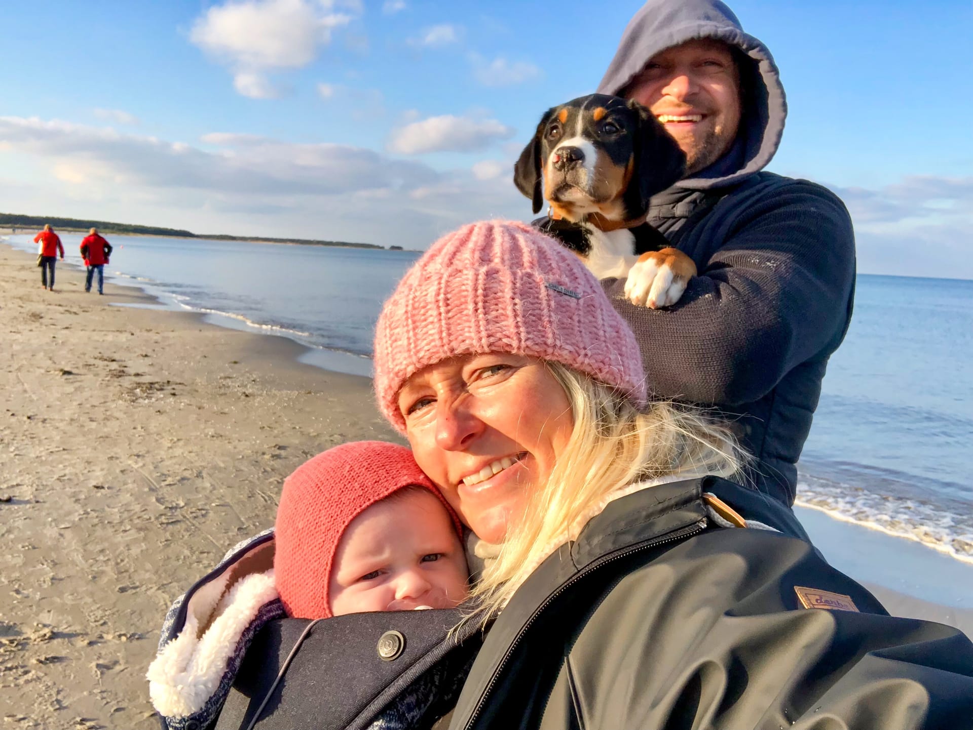 Familie am Strand: Frau mit Kind auf dem Arm, Mann mit Hund auf den Armen.