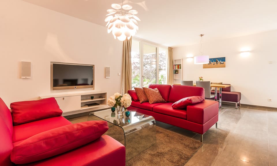 Ein Wohnzimmer mit zwei roten Ledersofas, einem Glastisch vor einem Wandfernseher und einem angrenzenden Essbereich.