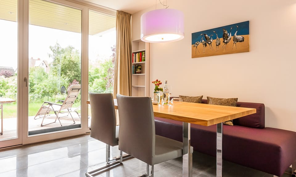 Ein Essbereich mit Holztisch, grauen Freischwinger-Stühlen und einer lila Sitzbank vor einem großen Terrassenfenster.