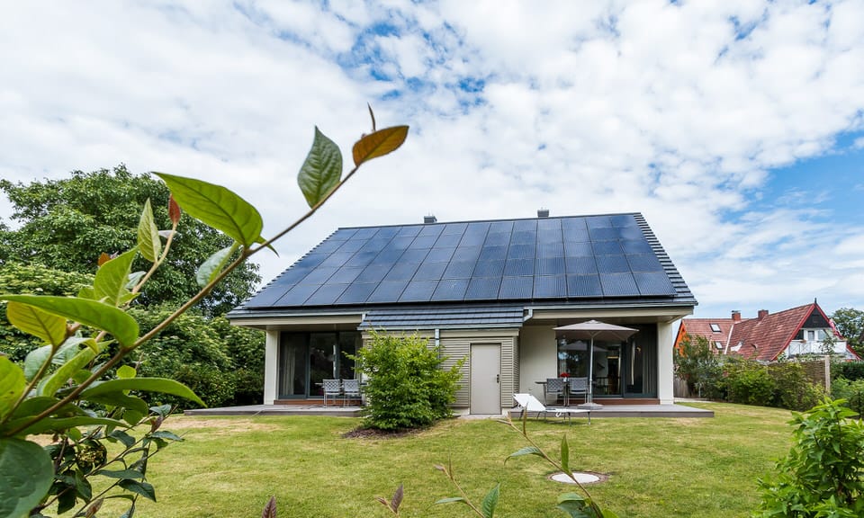 Ein Einfamilienhaus mit großer Photovoltaikanlage auf dem Dach und einem Garten mit Sitzmöbeln unter bewölktem Himmel.