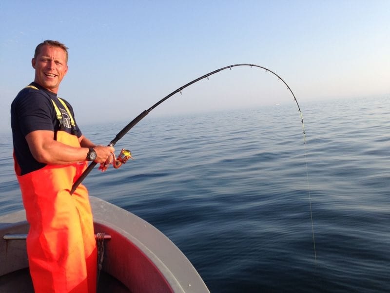 Ein lächelnder Mann auf einem Boot beim Angeln, dunkles T-Shirt und leuchtend orange Latzhose, hält eine gebogene Angelrute, ruhiges Meer und tiefstehende Sonne im Hintergrund.
