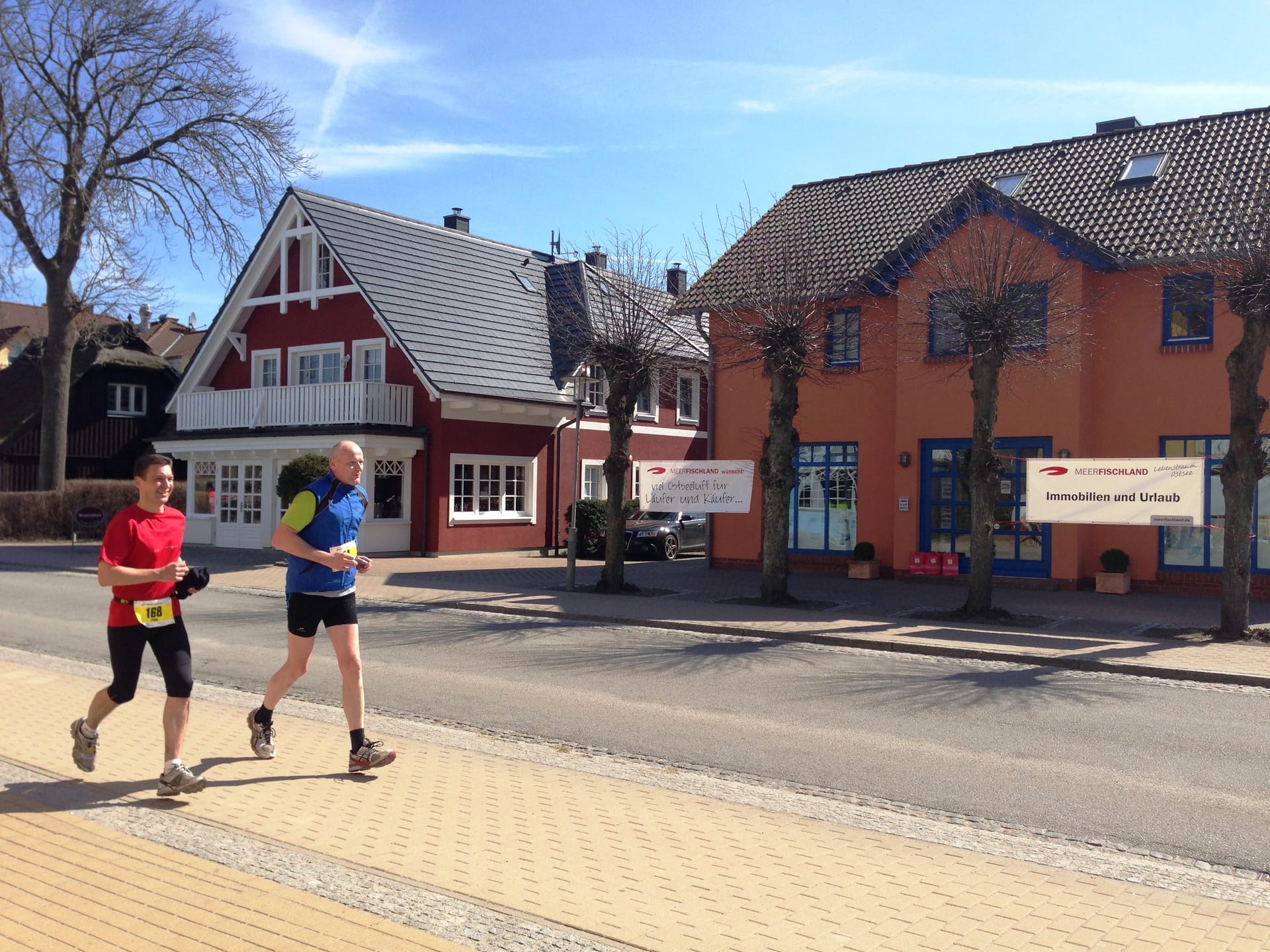 Zwei Läufer mit Startnummern auf einer sonnigen Straße vor einem roten und einem orangefarbenen Haus.