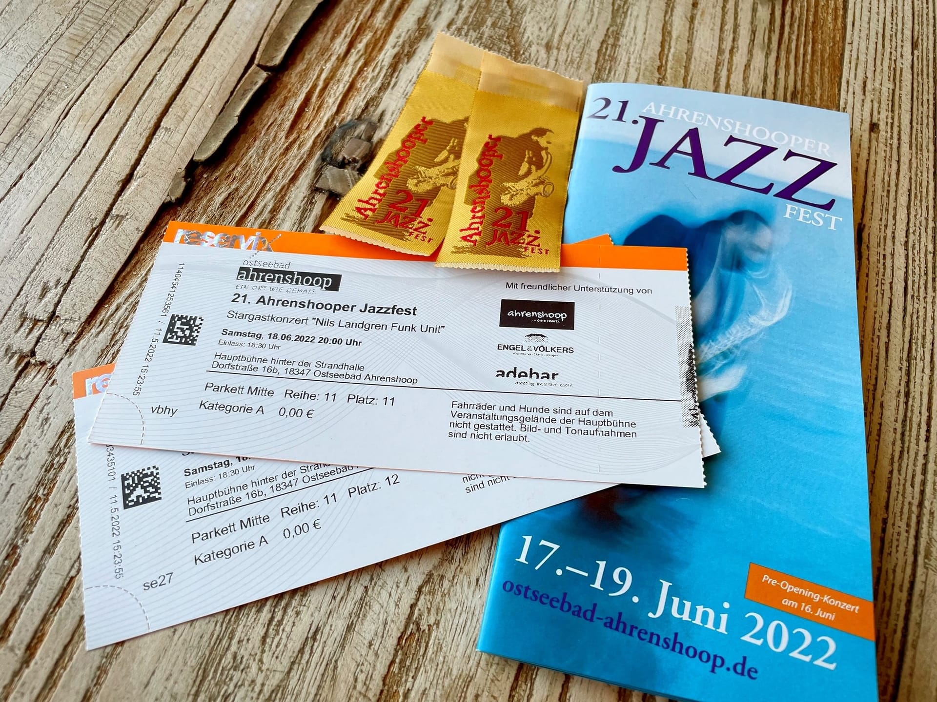 Zwei weiße Eintrittskarten für ein Konzert,  dahinter blaues Programmheft und gelbe Werbezettel auf hellem Holzhintergrund.