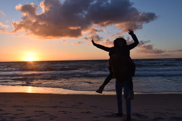 Zwei Personen sind am Strand. Eine Person trägt die andere auf dem Rücken. Die Sonne geht am Horizont unter. Das Meer ist im Hintergrund.