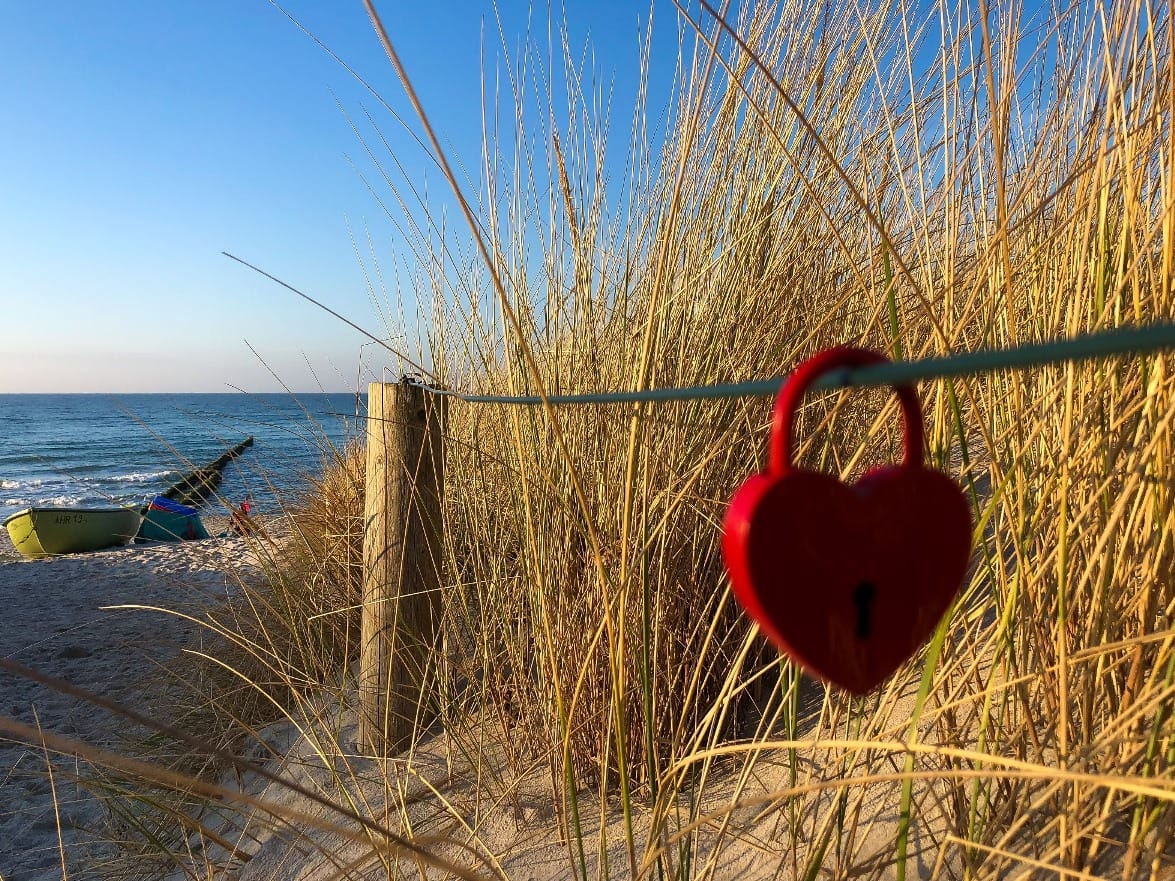 Ein rotes, herzförmiges Vorhängeschloss hängt an einem Drahtzaun am Eingang zum Strand.