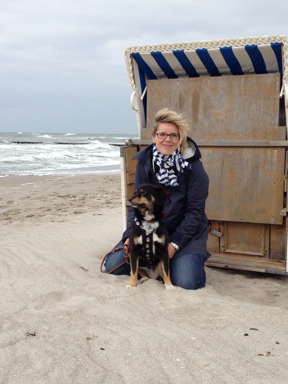 Eine Frau mit einem Hund sitzt am Strand neben einem Strandkorb.