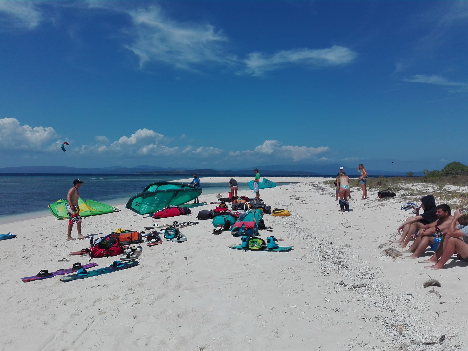 Mehrere Menschen halten sich an einem weißen Sandstrand mit türkisblauem Wasser auf, wo sie Kitesurf-Ausrüstung auslegen und sich sonnen.