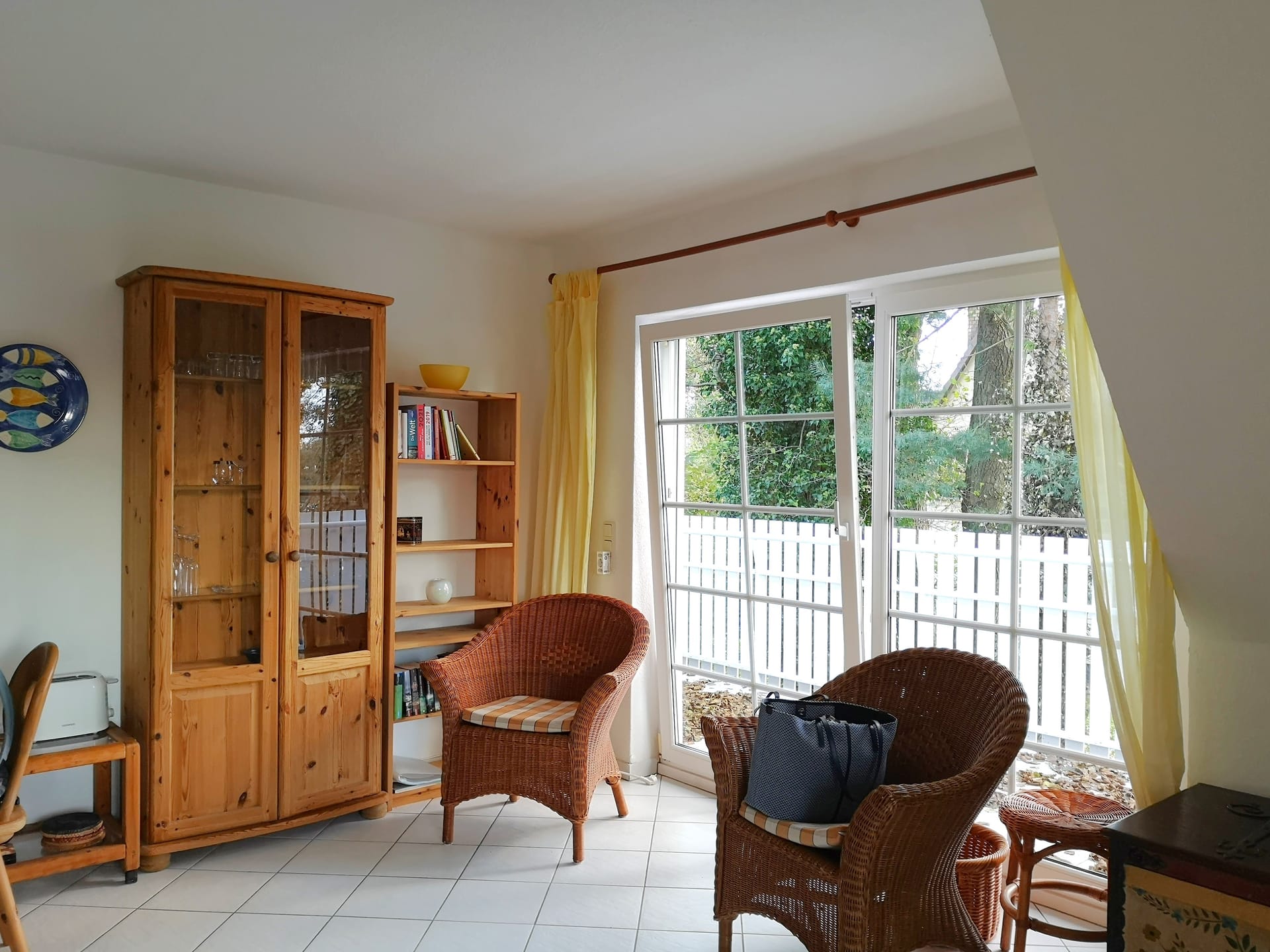 Ein Wohnraum mit zwei Korbsesseln, Holzschrank, Regal und großen Fenstern. Fliesenboden, Vorhänge und Blick in den Garten.