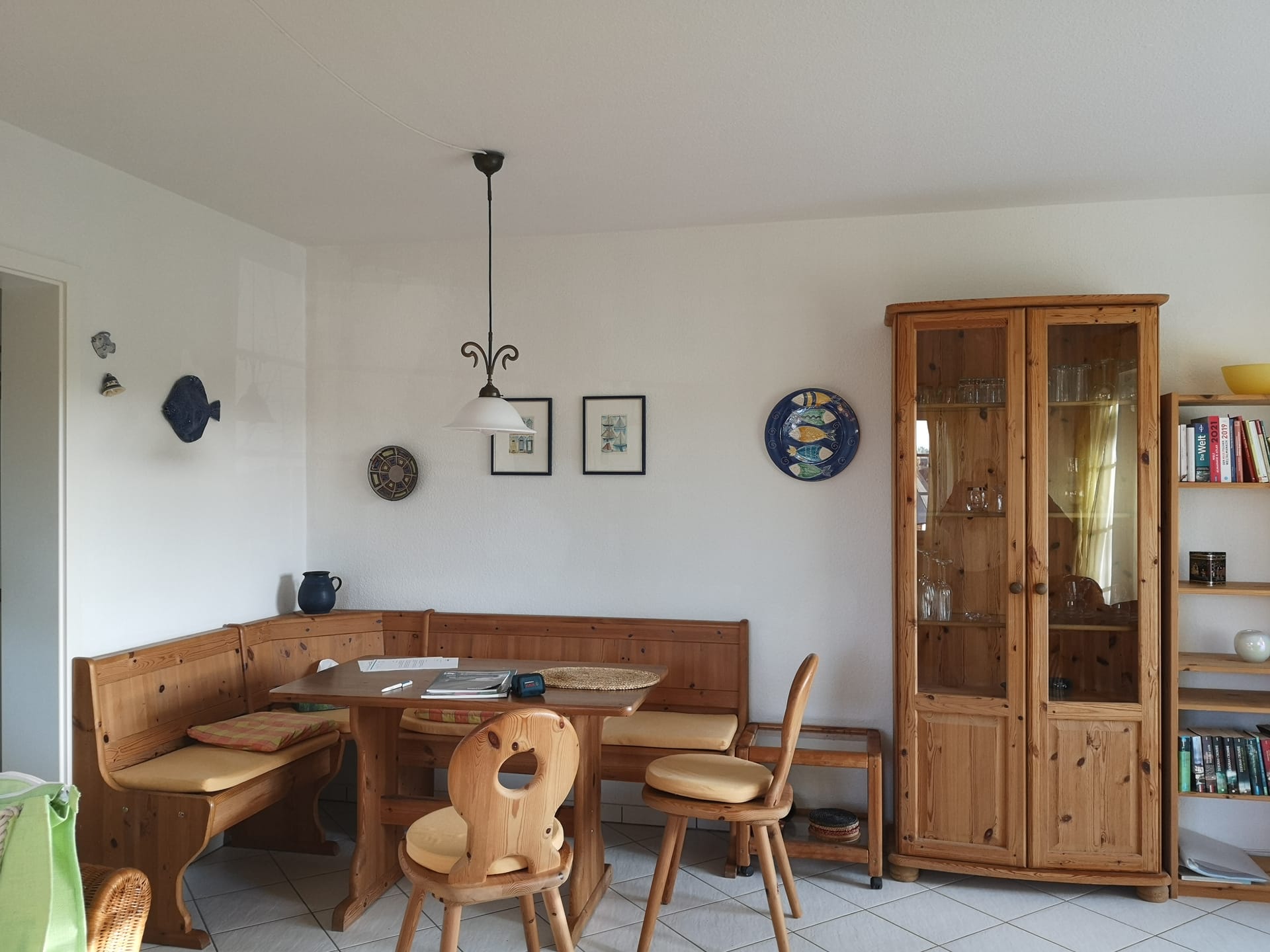 Der Essbereich ist mit Kiefernmöbeln eingerichtet. Es gibt eine Holzecke, einen Tisch und zwei Stühle. Rechts steht ein Vitrinenschrank im gleichen Kiefernstil, daneben ein Bücherregal.