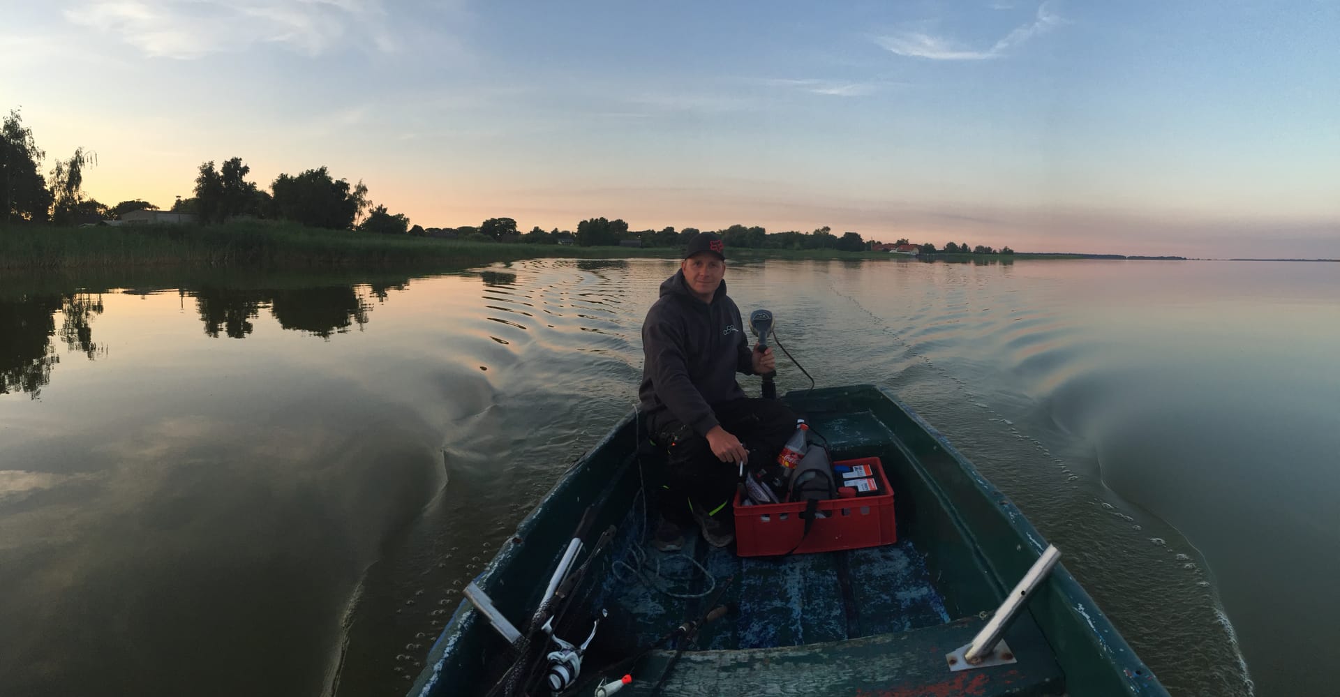 Ein Mann in dunkler Kleidung und Kappe in einem kleinen Boot auf ruhigem Wasser bei Sonnenuntergang, rote Kiste im Boot, flache Küstenlinie mit Bäumen im Hintergrund.