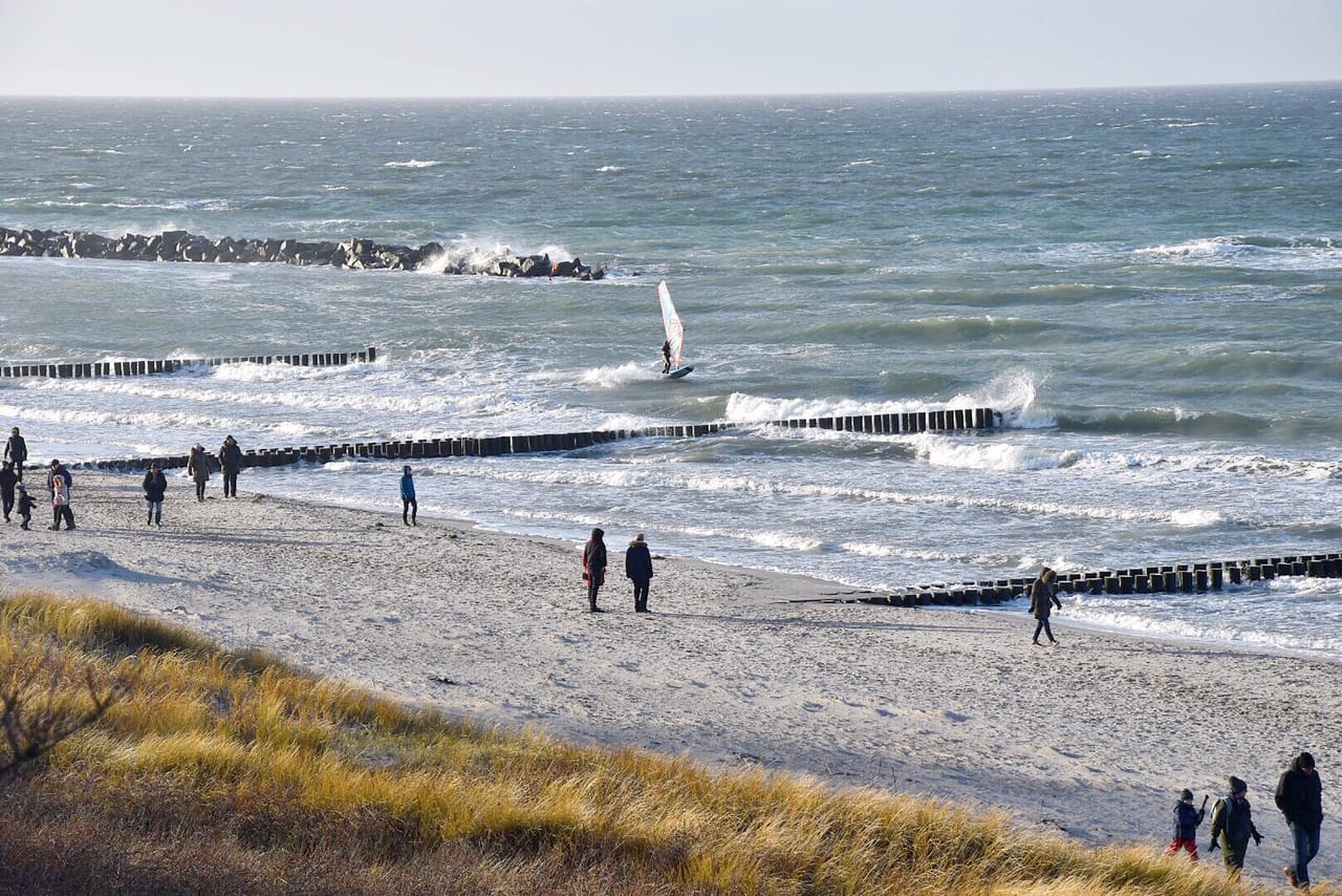 Menschen spazieren am Sandstrand. Im Meer sind Wellen, Buhnen und eine Person beim Windsurfen zu sehen.