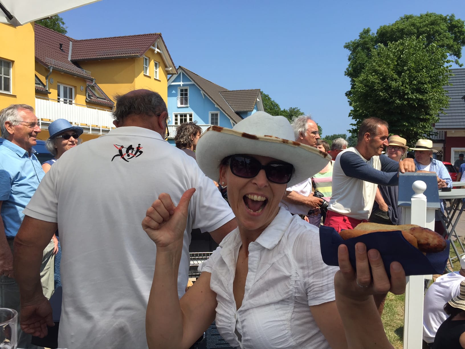 Eine lächelnde Frau mit Sonnenbrille und weißem Hut zeigt den Daumen nach oben und hält ein Brötchen mit Bratwurst.