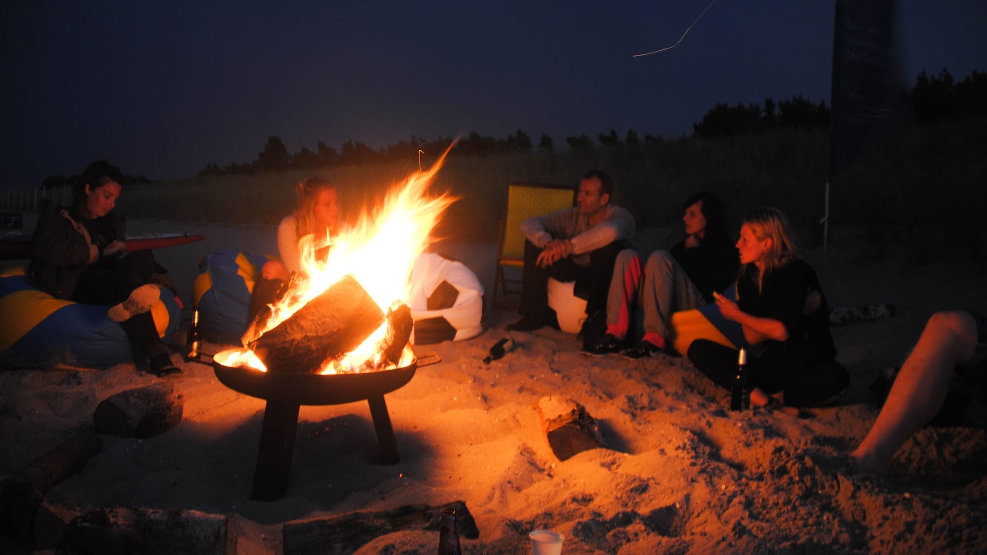 Mehrere Menschen sitzen nachts an einem Strand um eine Feuerstelle.