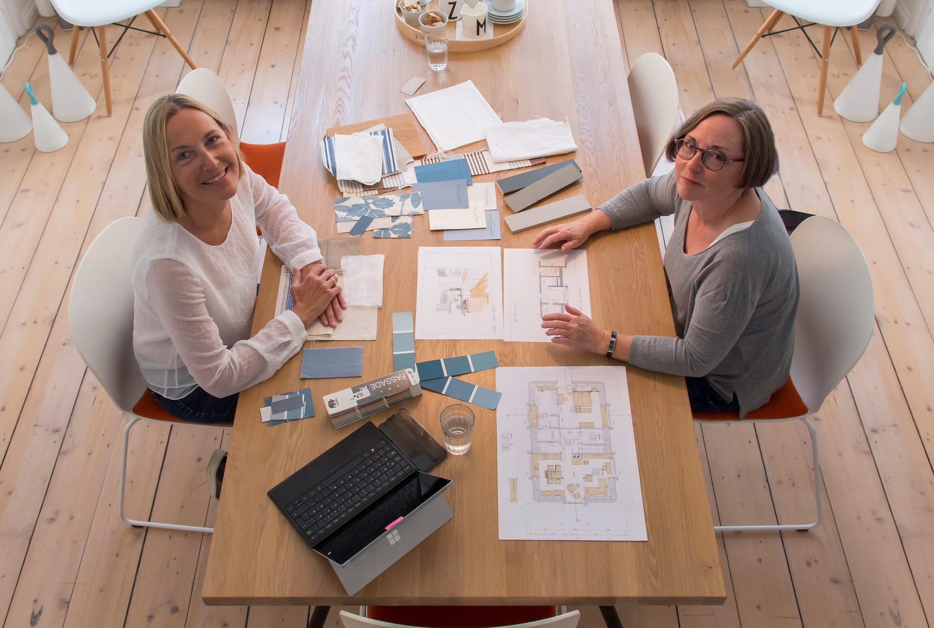 Zwei Frauen sitzen an einem großen Holztisch. Auf dem Tisch stehen ein Laptop, viele Dokumente, Grundrisse und verschiedene Materialproben.
