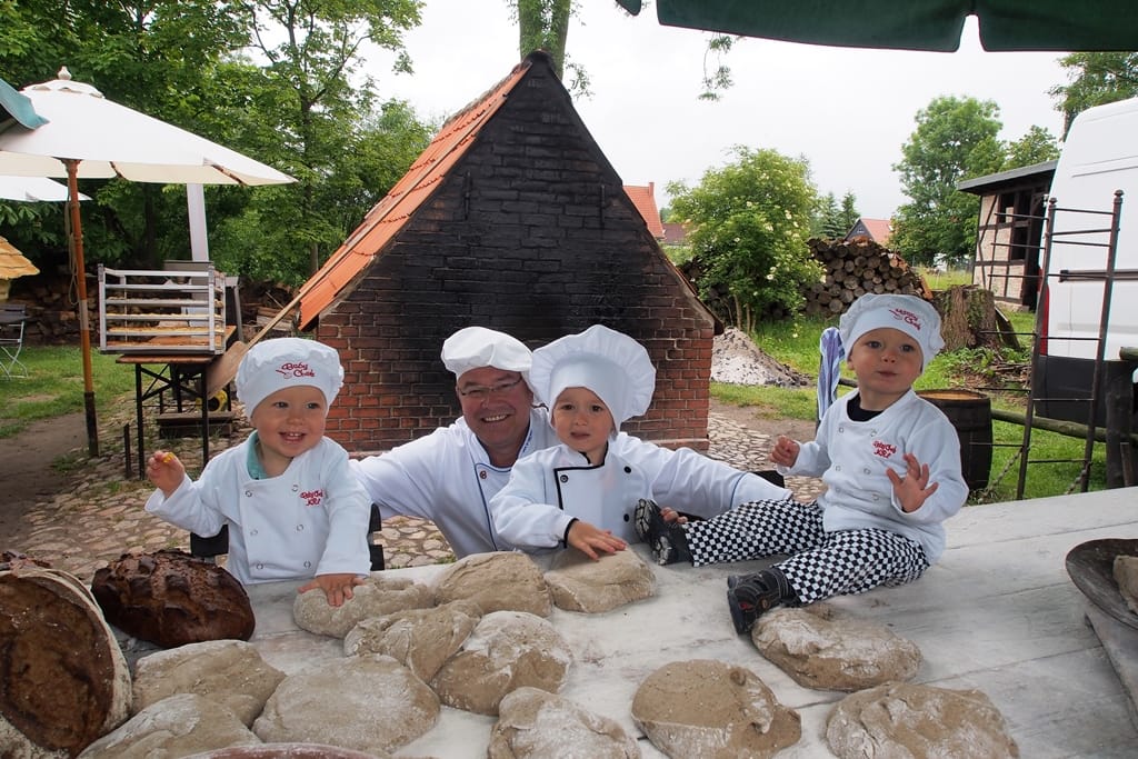 Eine Gruppe Kinder in Kochjacken und Kochmützen mit einem Bäcker am Tisch mit Brot und Brotteig.