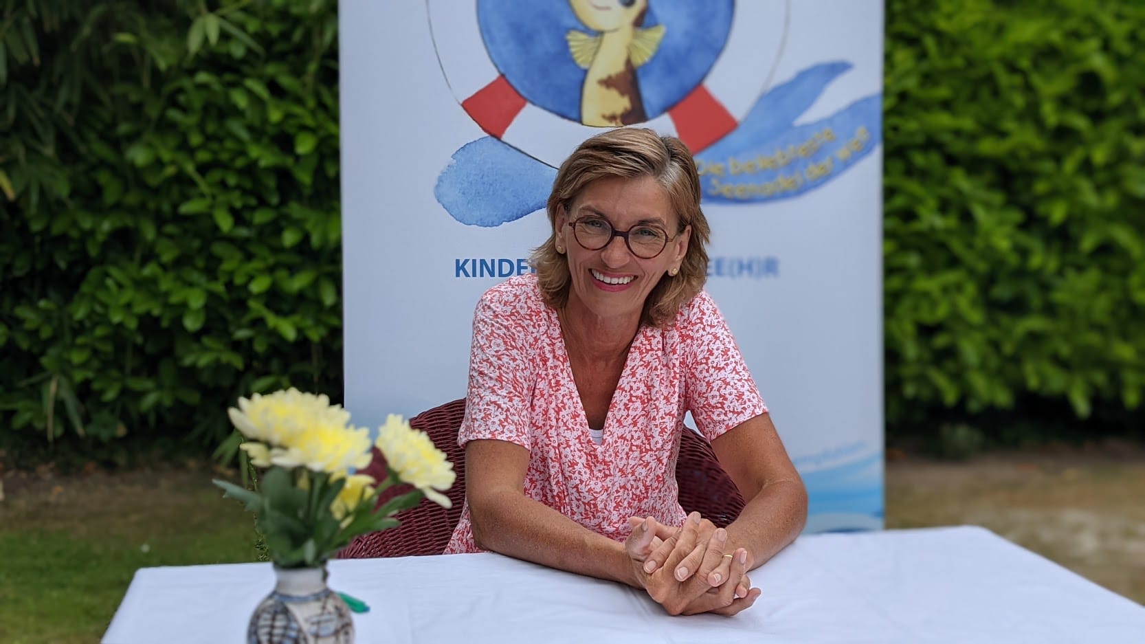 Eine lächelnde Frau mit Brille vor einem Plakat für Kinderbücher.