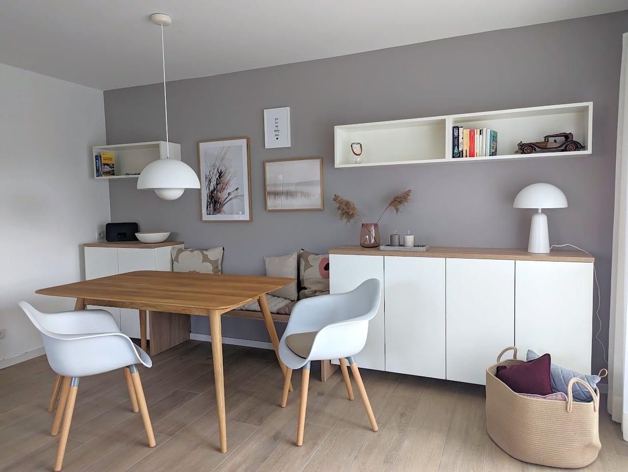Essbereich mit einem Holztisch, zwei weißen Stühlen, einer Sitzbank mit Kissen, einer weißen Kommode und einer grauen Wand mit Bildern. Holzboden und moderne, schlichte Einrichtung.