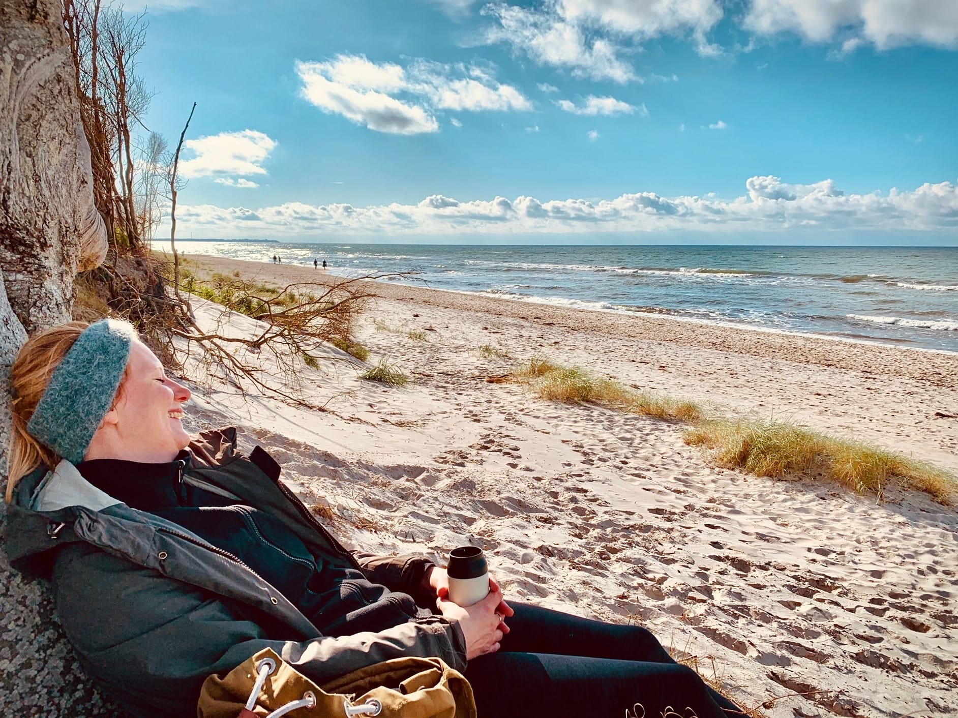 Eine Person liegt am Strand unter einem Baumstamm. Der Himmel ist blau mit weißen Wolken, und im Hintergrund ist das Meer zu sehen.
