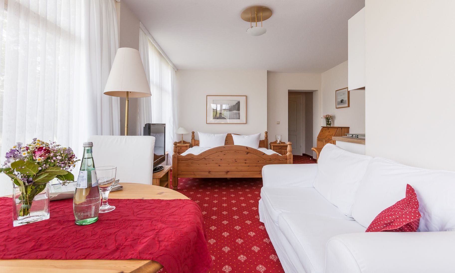 Innenansicht der Wohnung: Links ein Tisch mit Blumen darauf, ein Stuhl am Tisch, dahinter eine Lampe, weiter hinten ein Fernseher. Rechts ein Sofa. Hinten ein Doppelbett mit zwei Nachttischen, auf einem eine Lampe.