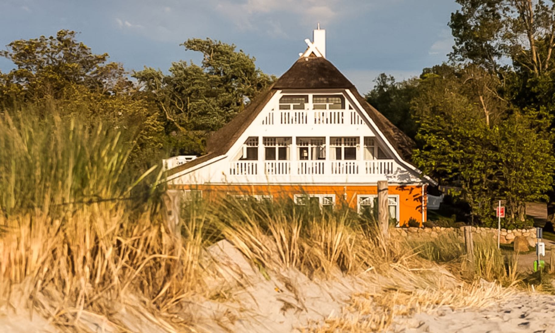 Ein orangefarbenes Haus mit Reetdach und weißen Balkonen, aus der Perspektive der Dünen gesehen.