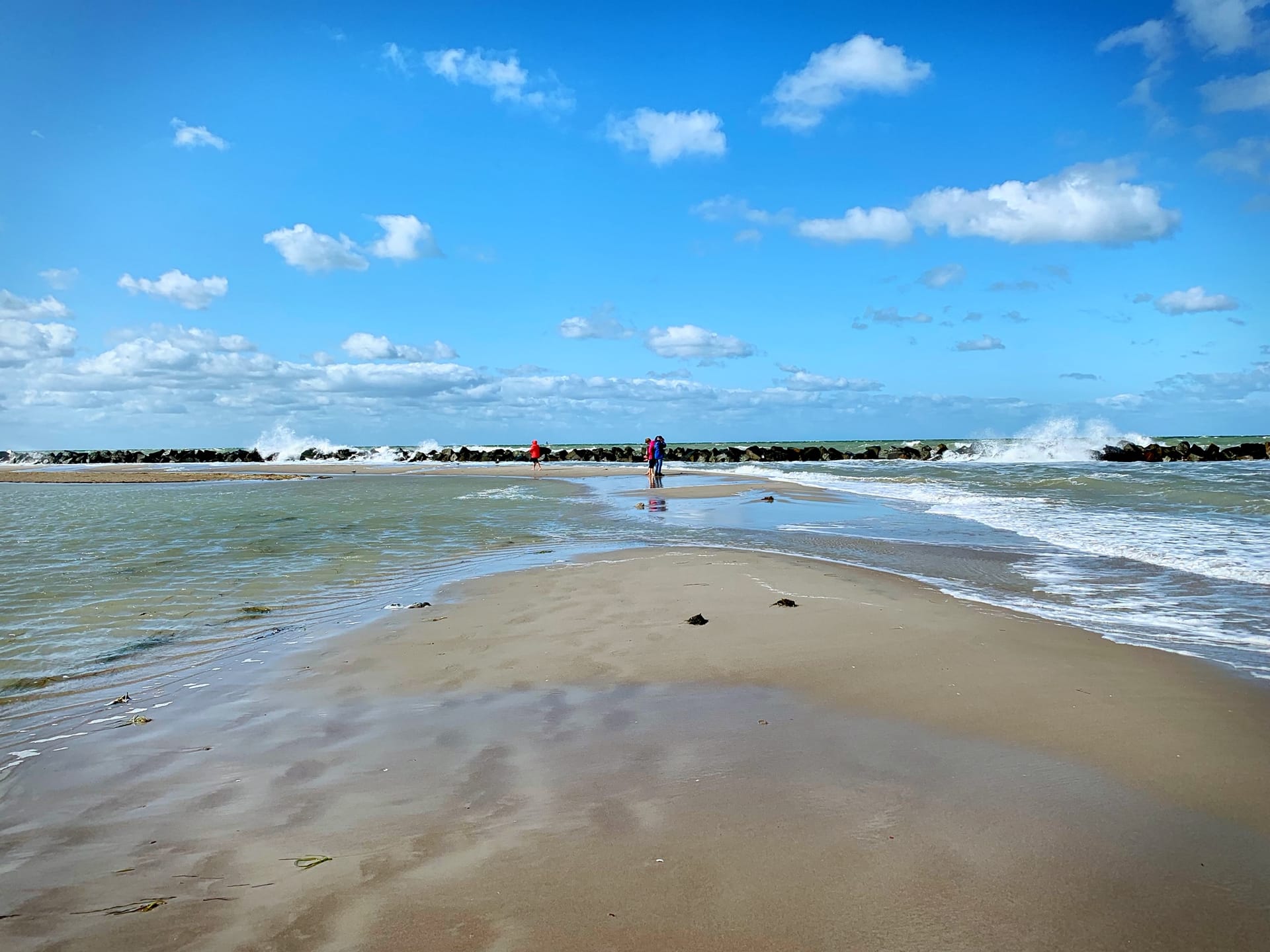 Wellen schlagen gegen den Wellenbrecher, einige Menschen am Strand, blauer Himmel.