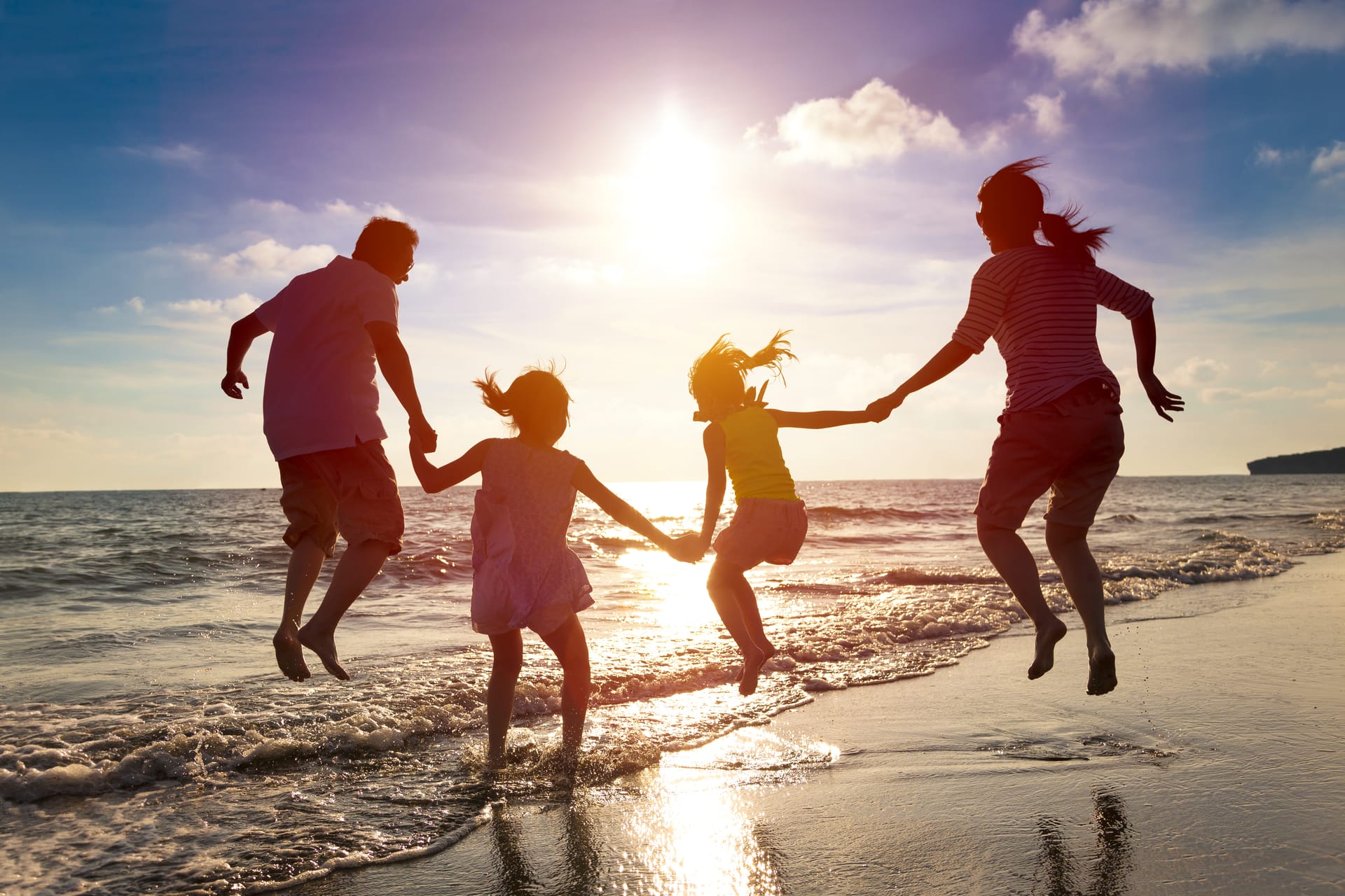 Zwei Erwachsene mit zwei Kindern springen am Strandufer Hand in Hand bei Sonnenuntergang.