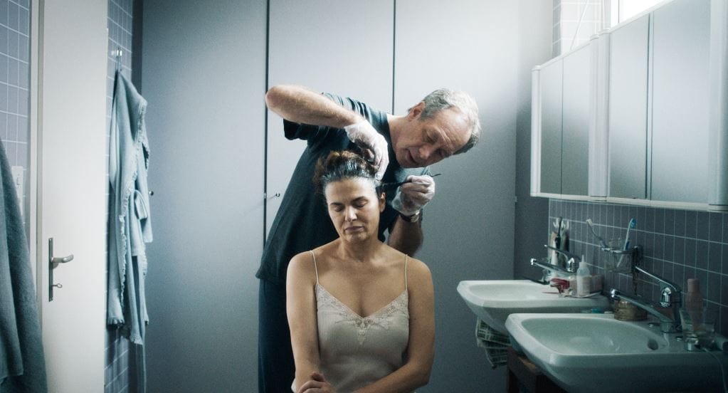 In einem Badezimmer eine sitzende Frau und ein hinter ihr stehender Mann beim Färben ihrer Haare, der Mann mit Handschuhen.