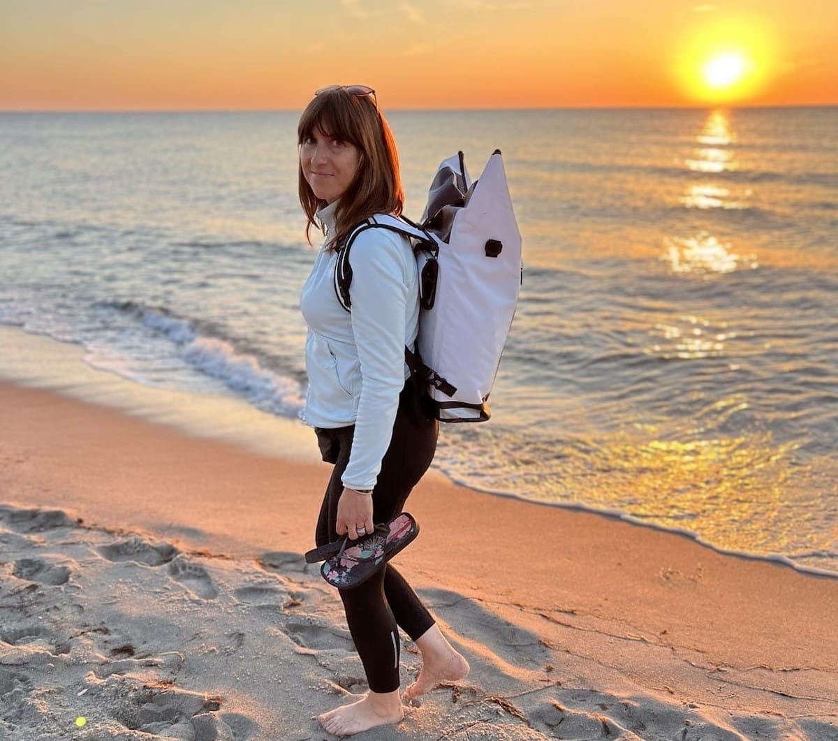 Eine Person mit einem Rucksack steht am Strand. Im Hintergrund ist der Sonnenuntergang sichtbar.