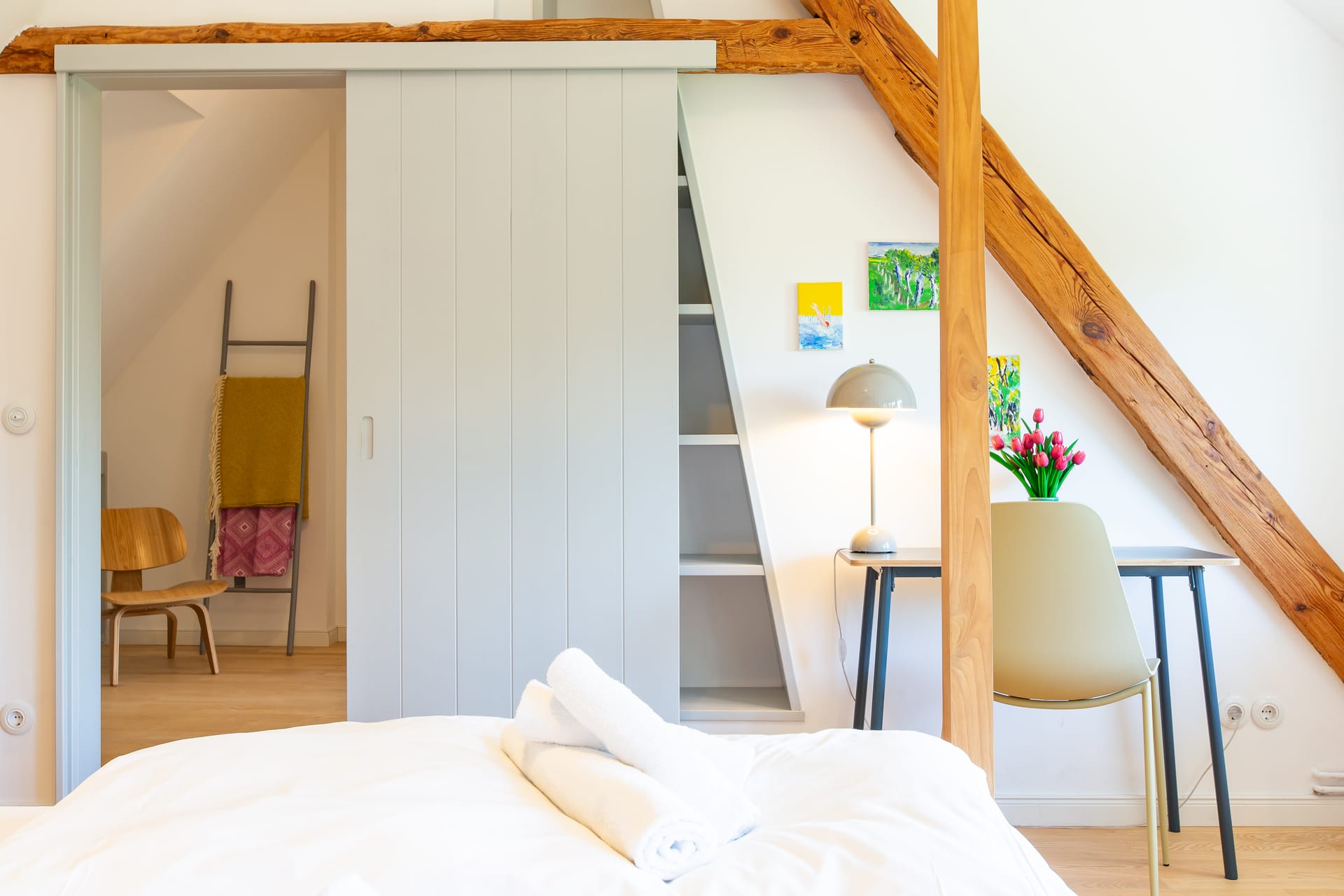 Ein Teil des Bettes ist sichtbar, darauf sind zwei zusammengelegte Handtücher. Dahinter ein Schreibtisch mit Stuhl. An der Wand über dem Schreibtisch hängen bunte Bilder. Das Schlafzimmer wird durch weiße Schiebetüren abgeschlossen.