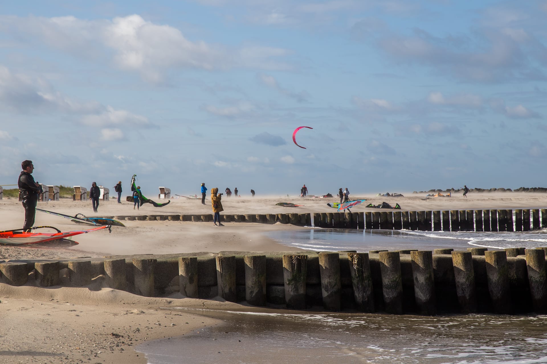 Ein Strand mit Holzpfählen zeigt Menschen beim Kitesurfen und Windsurfen auf dem Wasser unter bewölktem Himmel.