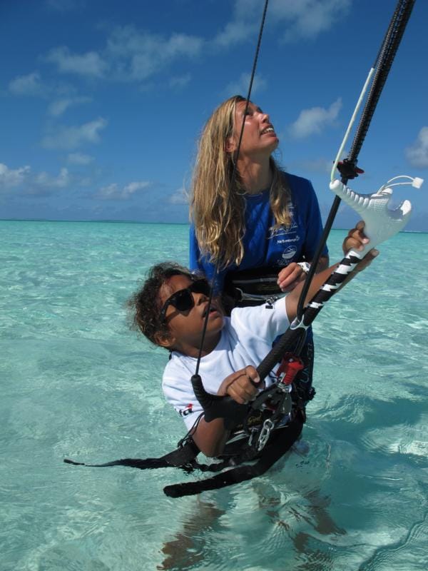 Zwei Personen tragen Kitesurf-Ausrüstung und blicken in klarem, blauem Wasser nach oben.