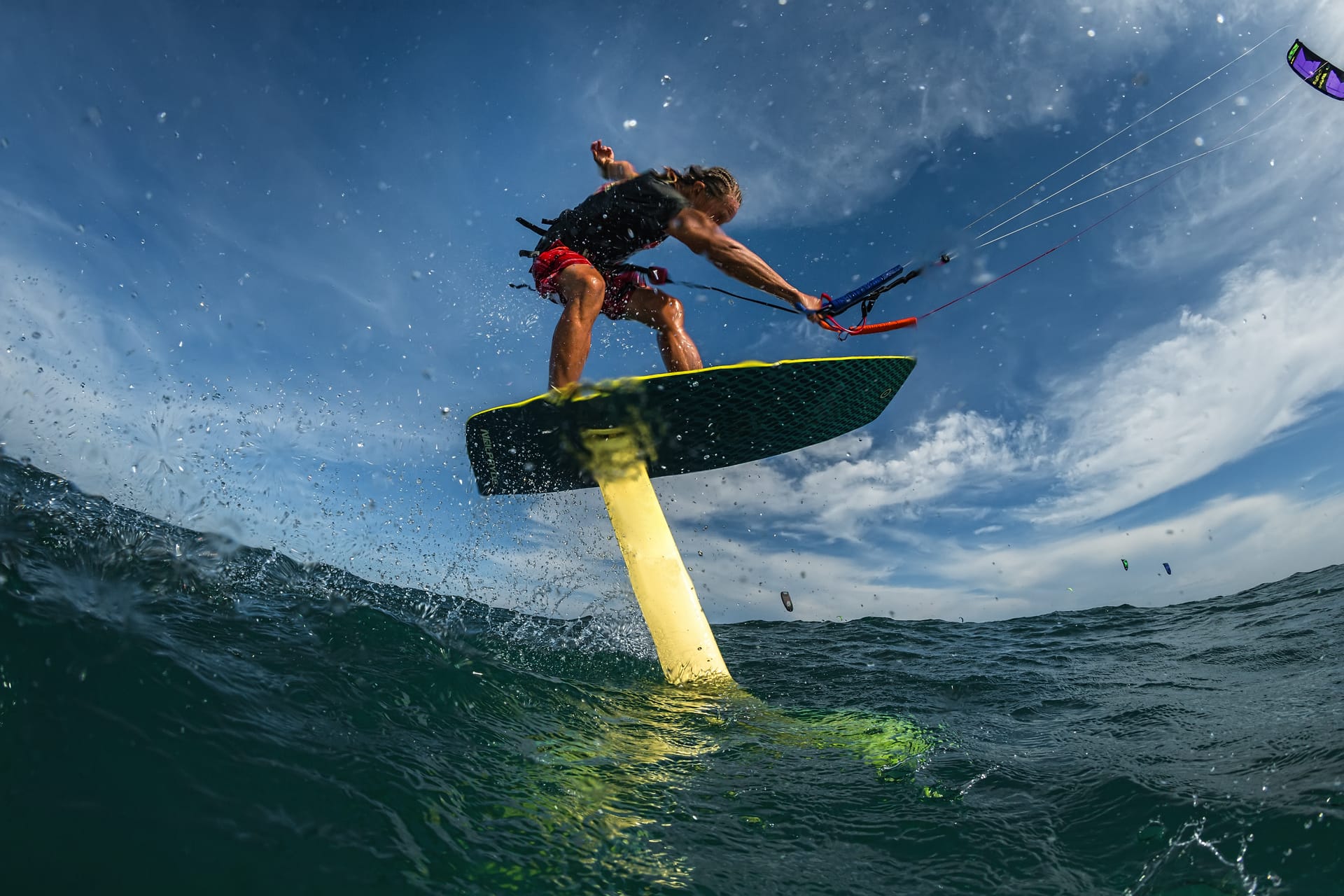 Eine Person steht auf einem Surfbrett mit Hydrofoil und betreibt Kitesurfen auf dem Wasser unter blauem Himmel mit Wolken.