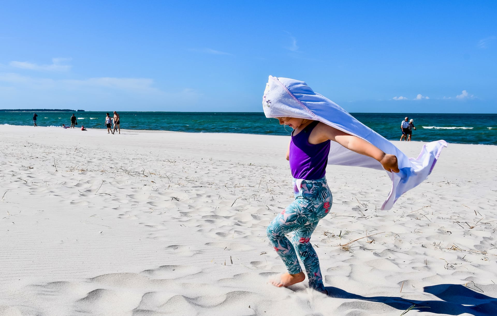 Kind läuft barfuß über den Sandstrand, mit Handtuch wie ein Cape, Ostsee im Hintergrund.