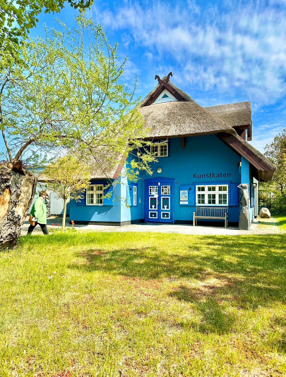 Blaues Reetdachhaus „Kunstkaten“ in Ahrenshoop, umgeben von grünem Garten.