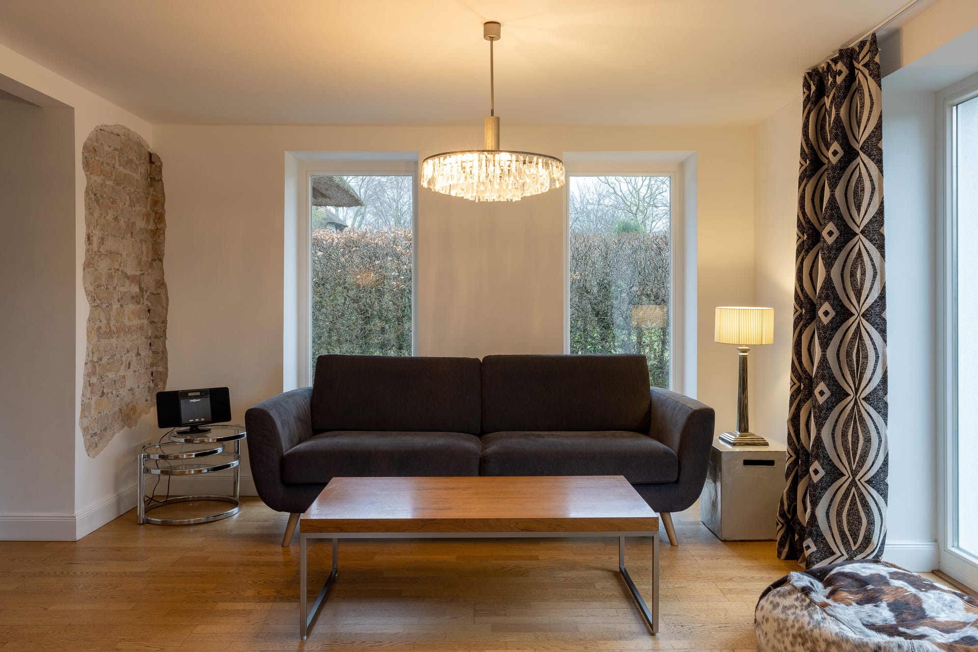 Wohnzimmer mit dunkelgrauem Sofa, Holztisch, Kronleuchter und einer freigelegten Steinwand an der Seite.